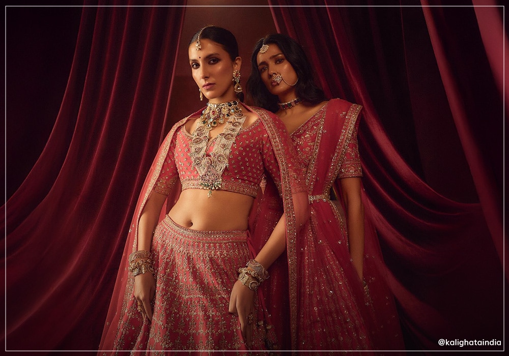 Two models wearing Kalighata outfits. One of the left wearing a red lehenga choli and one at the back wearing a red saree
