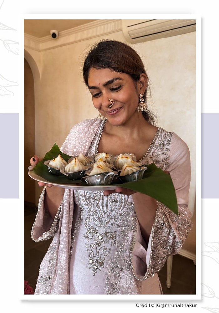 Mrunal Thakur wearing a shimmering silver kurta and dupatta and holding a plate of modak