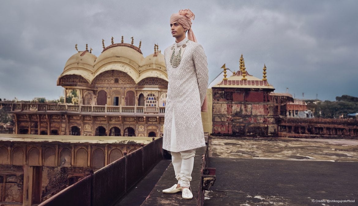 Groom wearing an ivory embroidered sherwani paired with a peach turban, churidar pants, and layered emerald necklace, photographed against a majestic Indian palace backdrop.