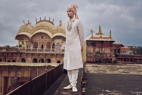Groom wearing an ivory embroidered sherwani paired with a peach turban, churidar pants, and layered emerald necklace, photographed against a majestic Indian palace backdrop.