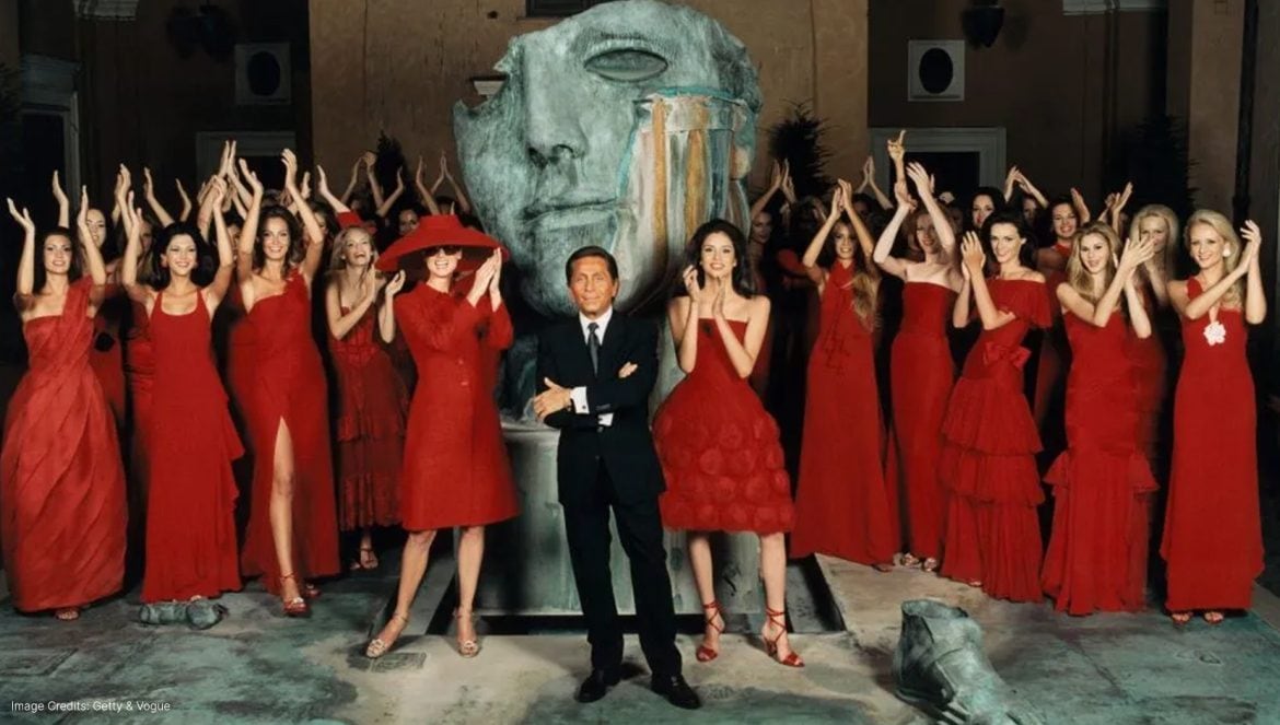 Models dressed in iconic red couture gowns stand in formation around a legendary fashion designer