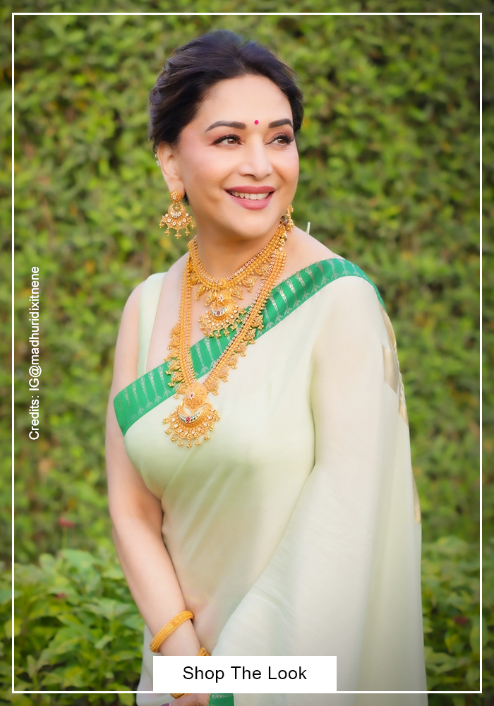Madhuri Dixit Nene in a close-up portrait wearing a House of Masaba half-and-half sari in pale mint with a green and gold border, hot pink printed lower drape peeking through, styled with a layered gold haar, gold bangles, chandelier jhumkas, and a red bindi against a green foliage background
