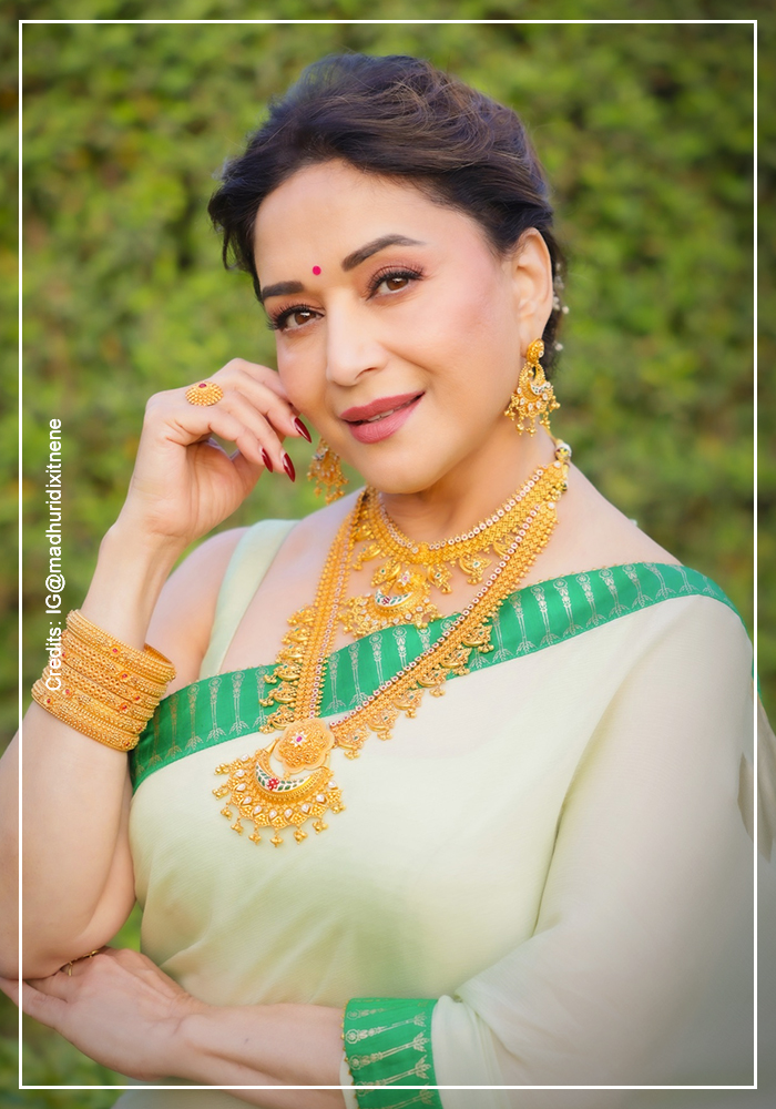 Madhuri Dixit Nene in a close-up portrait wearing a House of Masaba half-and-half sari in pale mint with a green and gold border, hot pink printed lower drape peeking through, styled with a layered gold haar, gold bangles, chandelier jhumkas, and a red bindi against a green foliage background